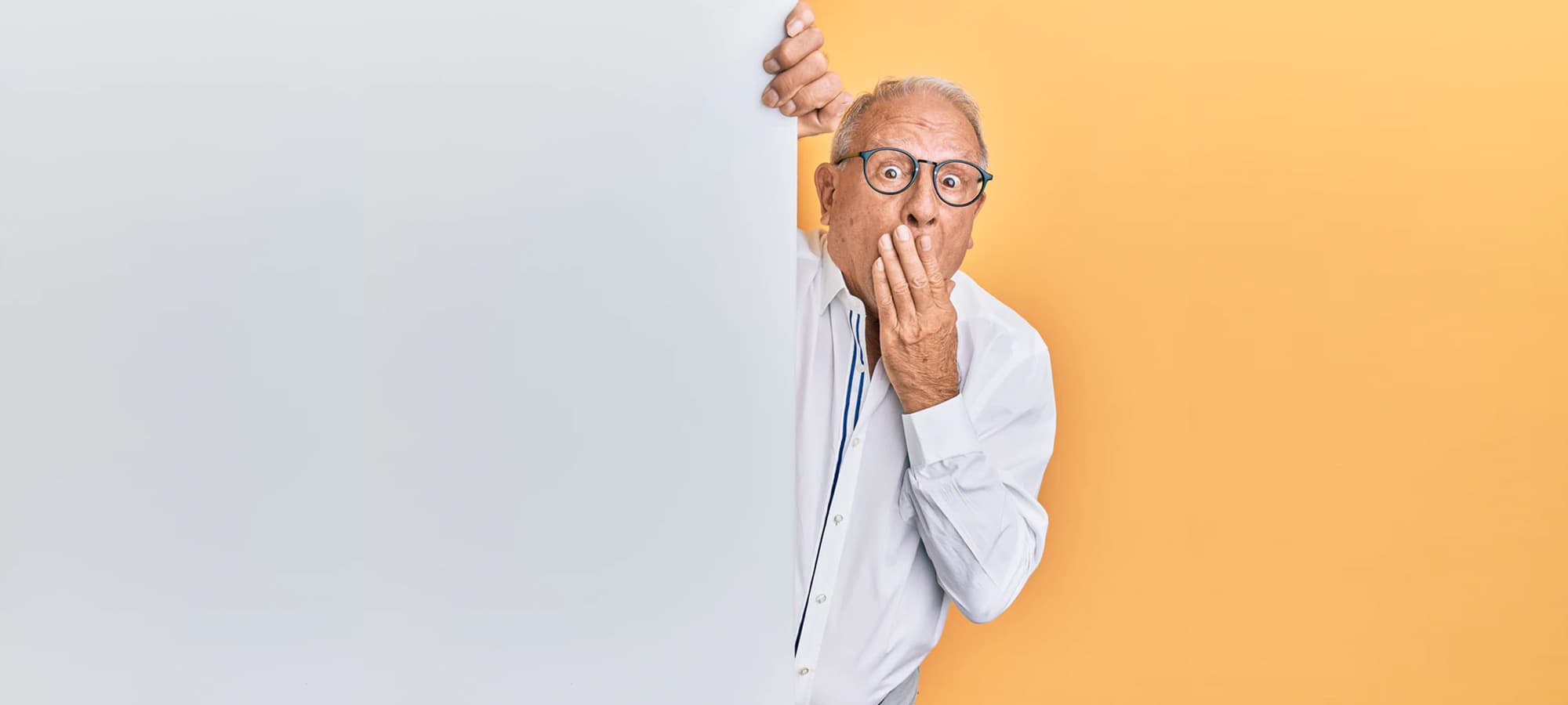 An elderly man wearing glasses peeks out from behind a wal